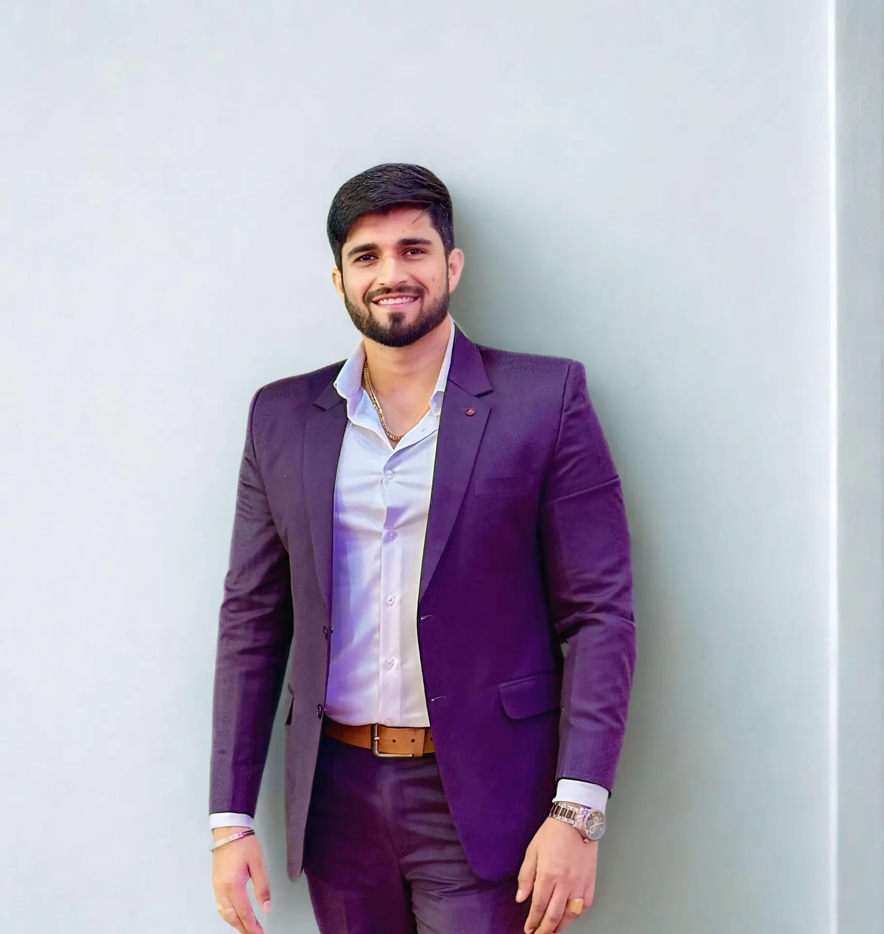 A man dressed in a formal suit, standing against a light-colored wall, smiling confidently.